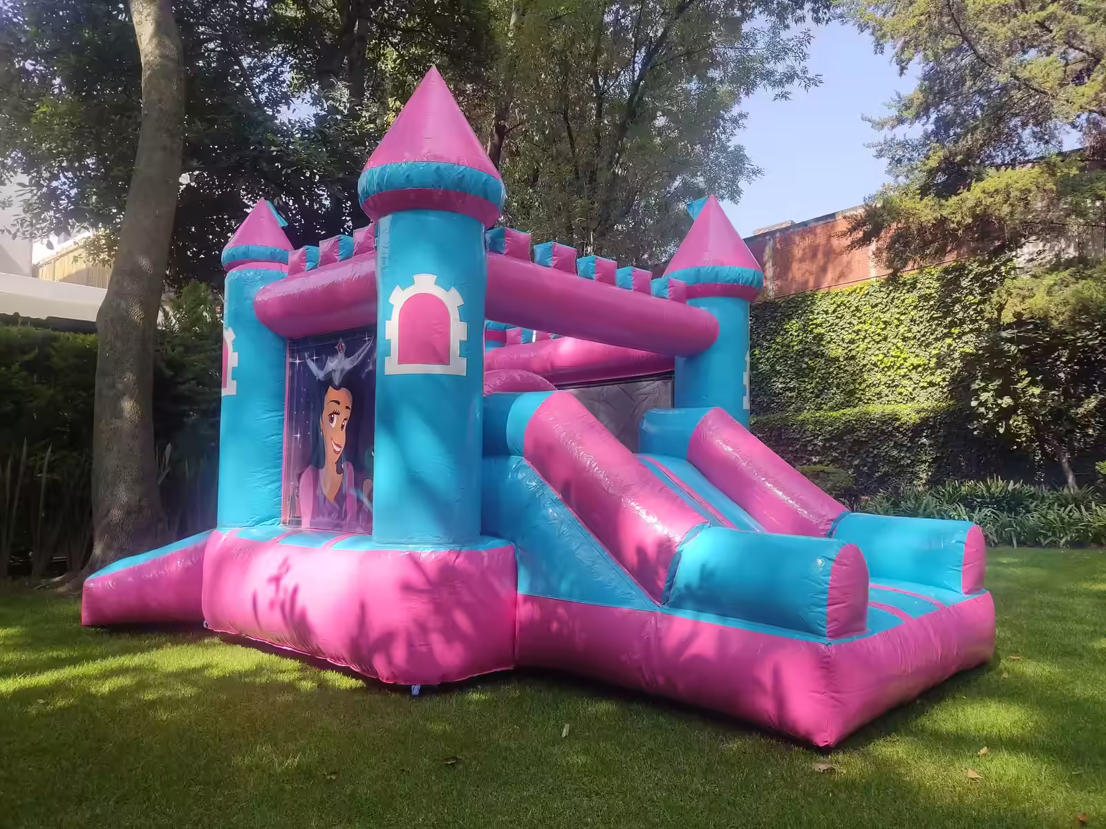 Inflable de castillo de princesas en tonos rosa y azul instalado en un jardín arbolado