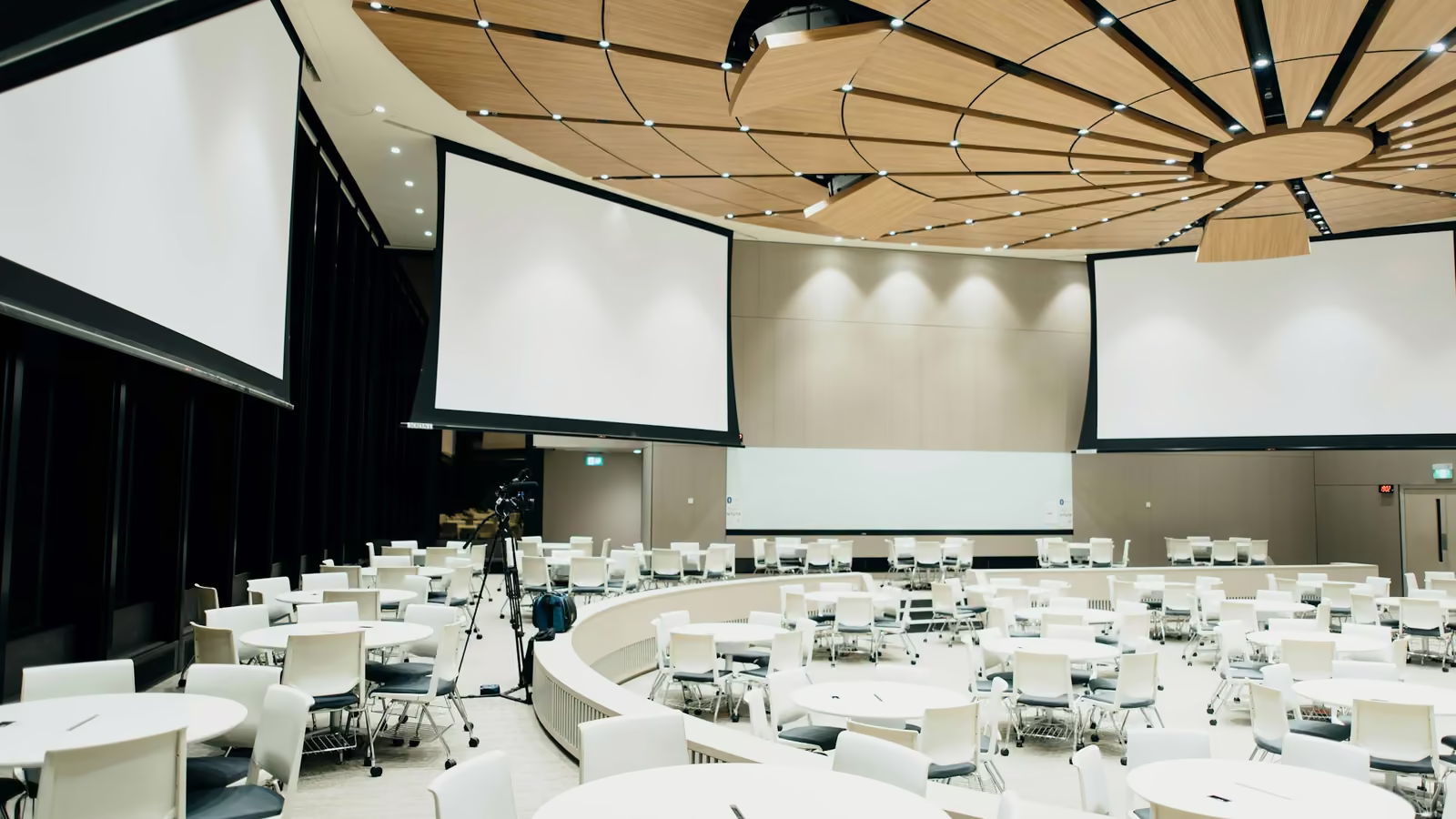 Sala de convenciones con mesas redondas, sillas blancas y tres pantallas de proyección
