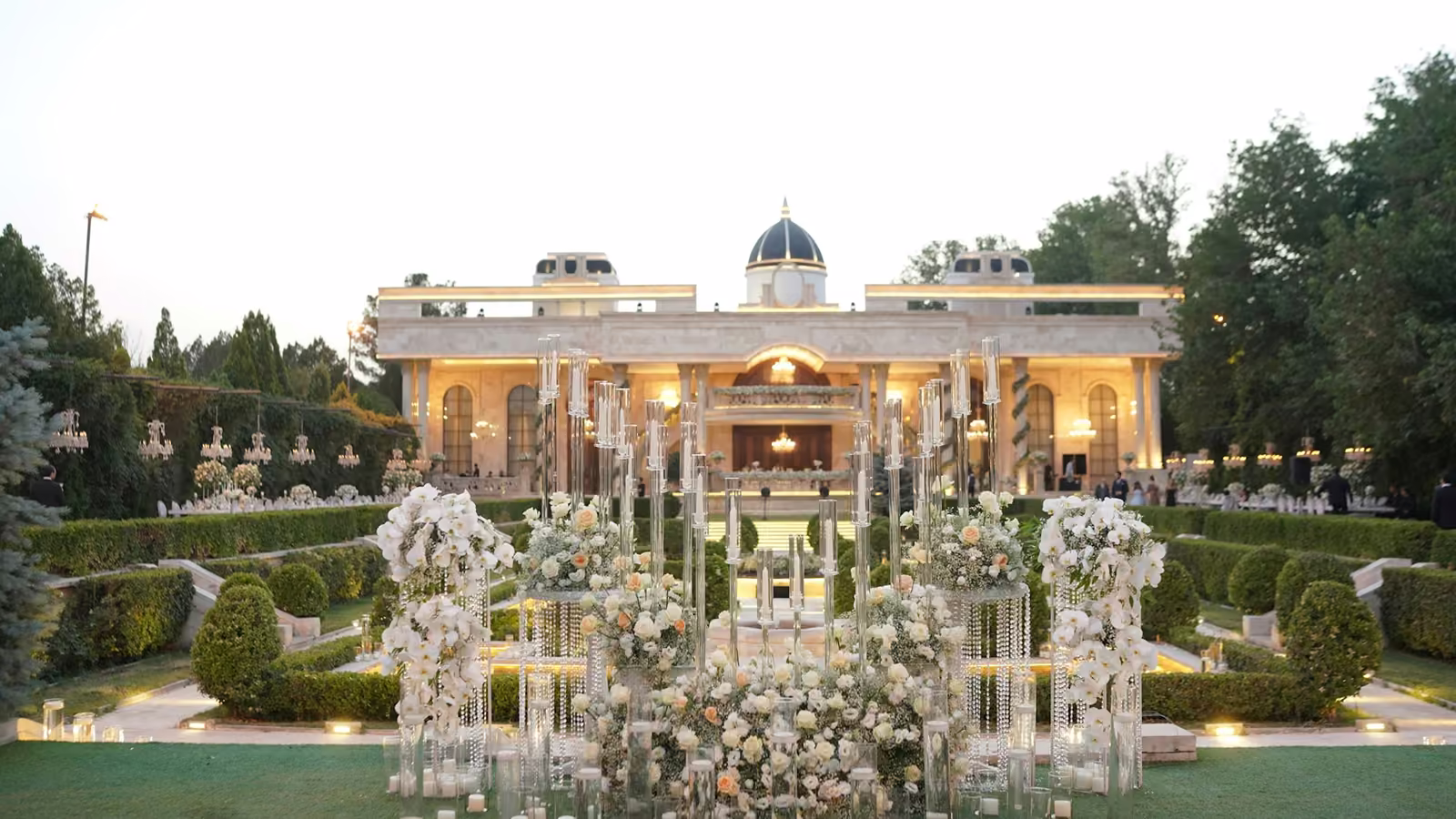 Arreglos florales y portavelas de cristal en un jardín frente a una casona